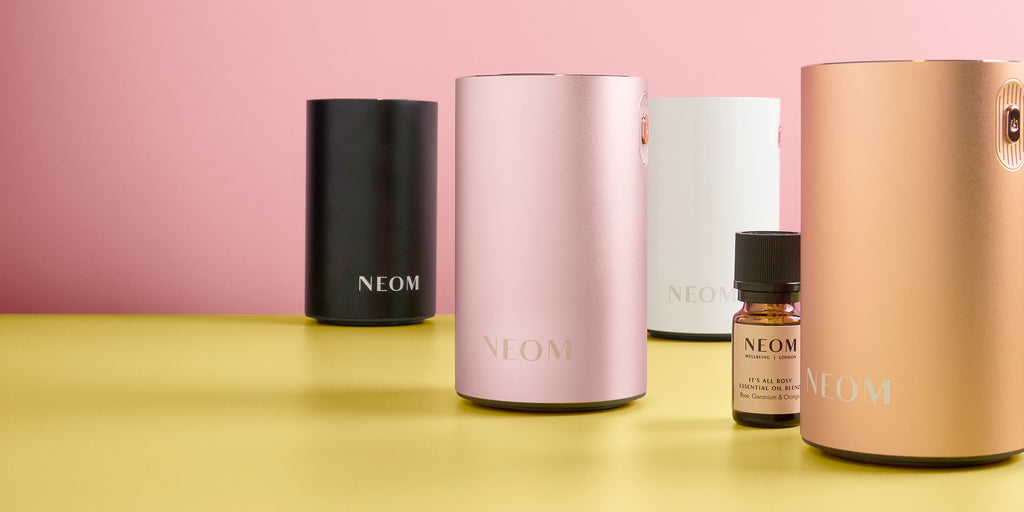 NEOM product lineup including a black, white, pink and gold Pod Mini+, and a small essential oil bottle on a yellow surface with a pink background.