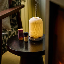 NEOM Electric pod diffuser on a small round table with bottles in a cosy room setting