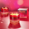 Two NEOM candles and their packaging on a red surface with a pink background