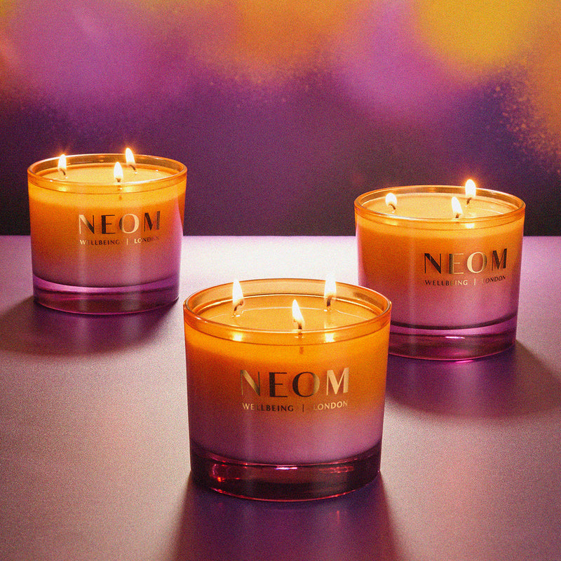 Three NEOM candles on a reflective surface with a blurred colorful background