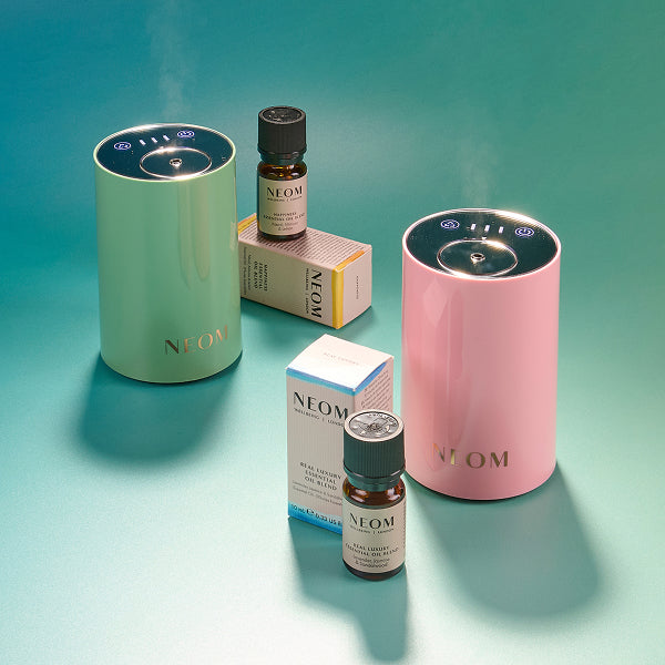 Two NEOM pod mini coloured diffusers, and essential oil bottles on a gradient blue background 