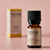 NEOM Happiness essential oil blend bottle with its box blurred in the background on a pink surface.