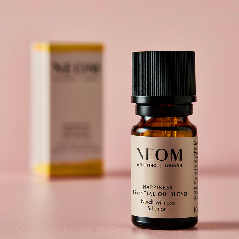 NEOM Happiness essential oil blend bottle with its box blurred in the background on a pink surface.
