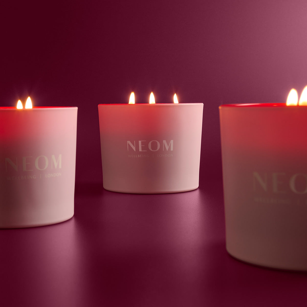 Three NEOM candles lit on a dark pink background