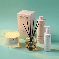 NEOM Wellbeing products including a candle, reed diffuser, and body cleanser on a gradient green background