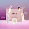 NEOM Wellbeing products including candles, bottles, and a box on a pink and purple gradient background.