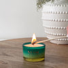 Lighting NEOM Perfect Peace candle in green glass jar on wooden table beside a white textured vase.
