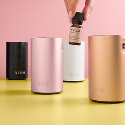 NEOM pod Mini+ collection including a bottle, pink, white, black and gold diffuser on a yellow surface with a pink background.