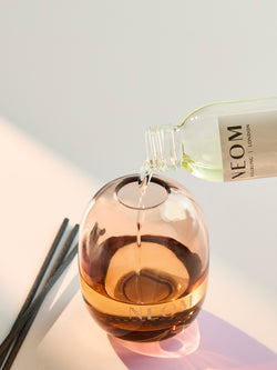 Pouring NEOM reed diffuser refill oil into glass diffuser bottle with black reeds on clean neutral background.